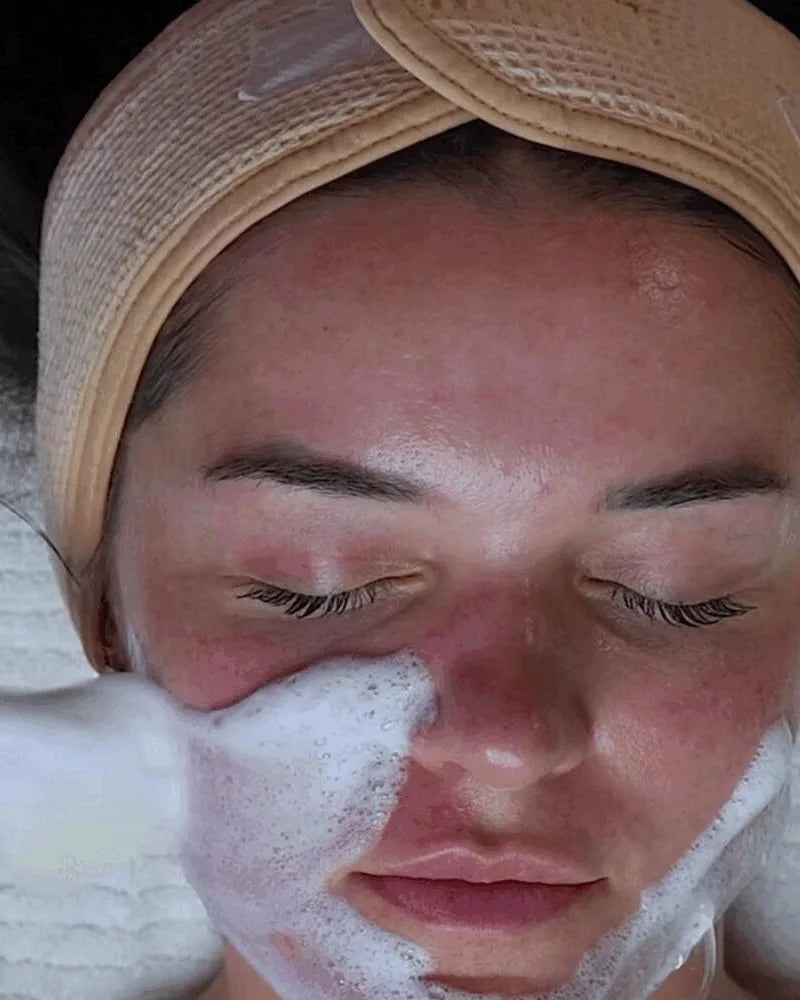 A guest resting with eyes closed under a headband as a soft white mask is brushed onto her cheek