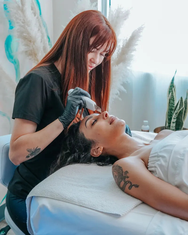 An esthetician in black gloves working a handheld skin tool along a resting guest's brow, pampas grass behind her