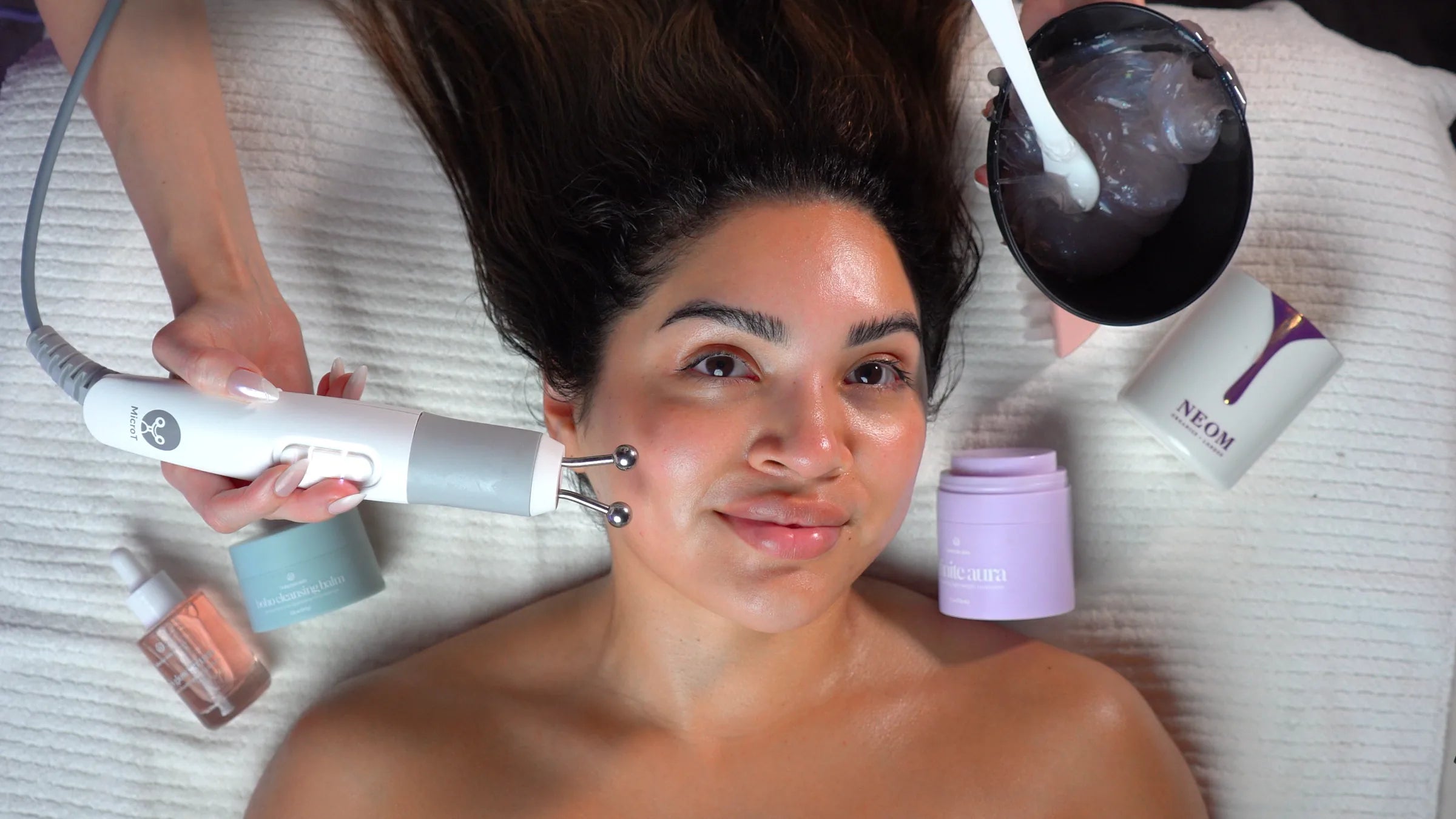 A smiling guest during a Préime Dermafacial, a microcurrent handpiece at her cheek and skincare laid out around her hair